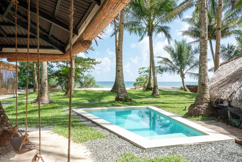A small rectangular pool surrounded by palm trees overlooks a beach, reminiscent of the tranquil scenes at cheap hotels on Siargao. A swing hangs from a thatched structure on the left, offering the perfect spot for relaxation.