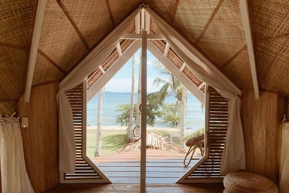 Interior view of a tropical bungalow on Siargao, featuring wooden walls and a triangular thatched roof. Through the open door, a deck with palm trees and a beach view invites you to explore this idyllic locale known for its charm over the cheap hotels nearby.