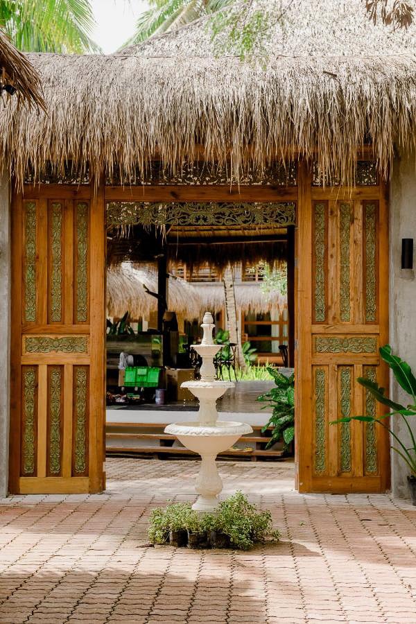 Open wooden doors lead to a tropical courtyard with a white fountain and greenery, surrounded by thatched roofs.