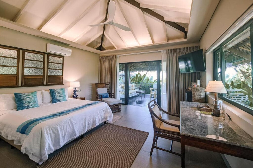 A well-lit hotel room with a double bed, desk, chair, and wall-mounted TV. Large glass doors open to a furnished patio surrounded by greenery. Ceiling fan and lamps provide lighting.