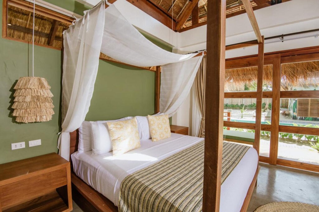 Canopy bed with white curtains, green accent wall, and wooden furniture. Large windows with bamboo shades. Brown rug on concrete floor. Natural light fills the room.