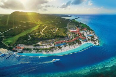 Aerial view of a coastal resort with buildings, beach, and clear blue water. A boat is sailing near the shore, and green hills are visible in the background. Discover the perfect destination at Sandals Resorts for your honeymoon.