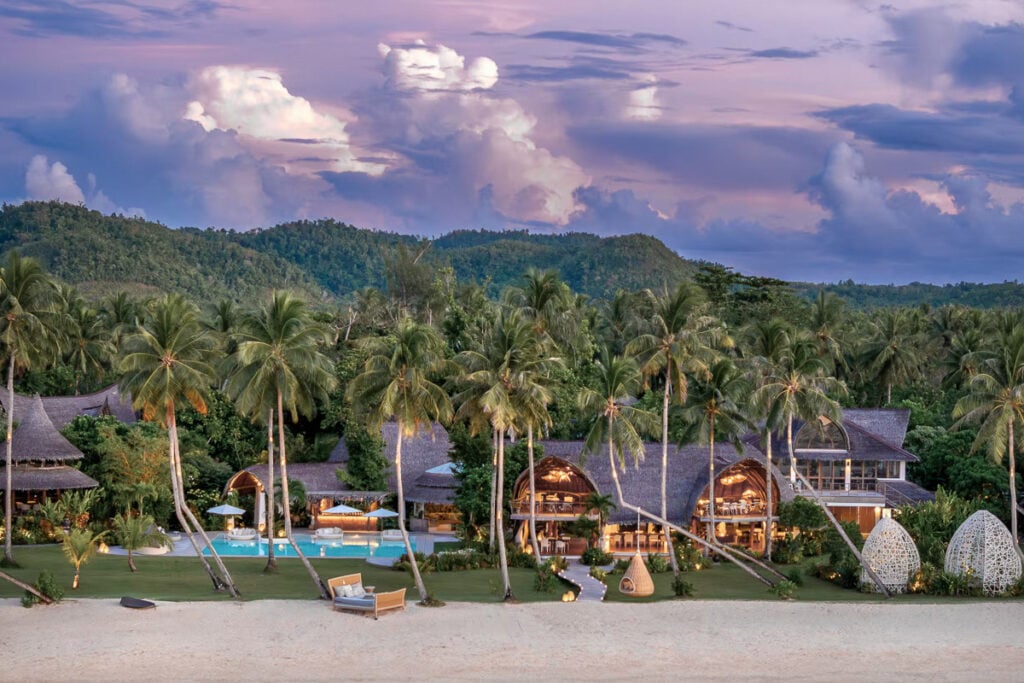 A beachfront resort in Siargao with villa-style buildings, palm trees, and a pool under a purple sky, surrounded by lush green hills. Discover the answer to "where to stay in Siargao" at this tropical paradise.